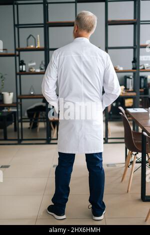 Back view of unrecognizable senior adult male doctor in white medical uniform standing in hospital office. Stock Photo