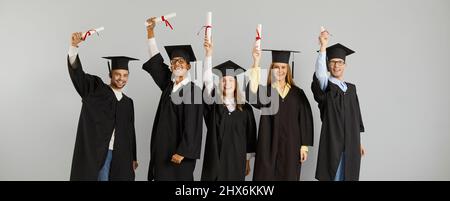 Joyful successful multiracial graduates with scrolls of diplomas in hands on gray background. Stock Photo