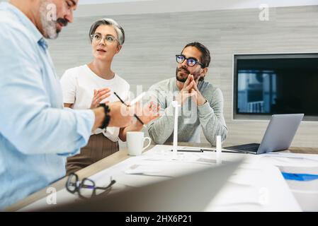 Group of design professionals having a discussion while working with wind turbine models and blueprints. Creative businesspeople working on a renewabl Stock Photo