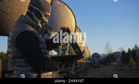 Hacker hacks the radio telescope using notebook. Man to program hacking on laptop in nature. Terrorist is hiding in camouflage balaclava. Hacker has criminal cases on the Internet. Programmer uses a computer to hack military communication technology. Stock Photo