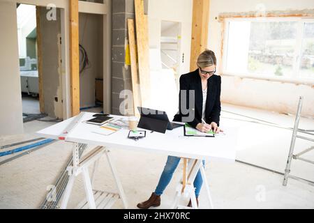 pretty young architect working on construction site in a loft and standing by a high table wearing glasses and a black jacket Stock Photo