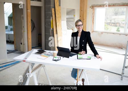 pretty young architect working on construction site in a loft and standing by a high table wearing glasses and a black jacket Stock Photo