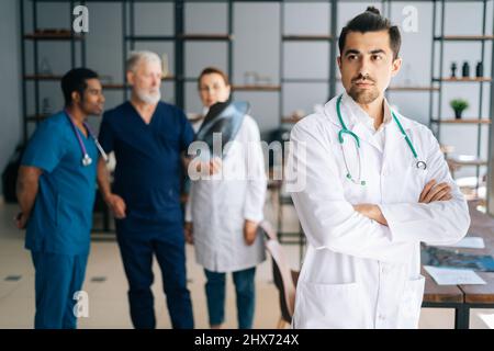 Portrait of confident young male doctor in white uniform standing with crossed hands in medical meeting office, looking away. Stock Photo