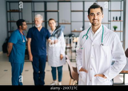 Portrait of cheerful young male doctor wearing white uniform standing in medical meeting office, looking at camera. Stock Photo