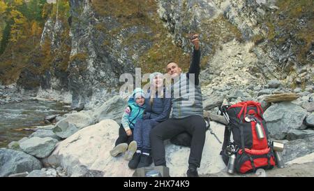 Tourists rest after hike and take selfie on the phone. Family travels. People environment by mountains, rivers, streams. Parents and kids walk using trekking poles. Man and woman have hiking backpacks. Dad mom and daughter sports travel family. Stock Photo