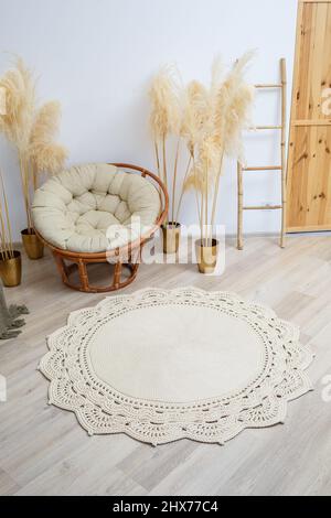 Homemade knitted rug made of thick threads, handmade: a handmade rug made of beige threads lies in the living room near the armchair, the art of knitt Stock Photo