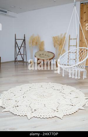 Homemade knitted rug made of thick threads, handmade: a handmade rug made of beige threads lies in the living room near the armchair, the art of knitt Stock Photo