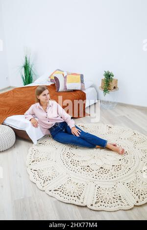 A woman lies on a rug near the bed, a homemade knitted carpet made of thick threads, made by hand: a handmade rug made of beige threads lies on the fl Stock Photo