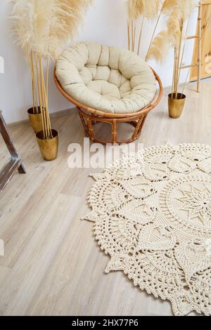 Homemade knitted rug made of thick threads, handmade: a handmade rug made of beige threads lies in the living room near the armchair, the art of knitt Stock Photo