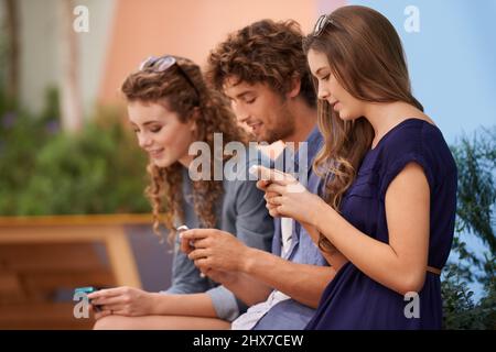 None of those awkward silent moments here.... Three young adults using their cellphones while sitting outdoors. Stock Photo