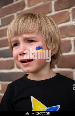 face of crying frightened boy, yellow-blue Ukrainian flag is painted on his cheek. Russia's invasion of Ukraine, request for help to world community. Stock Photo