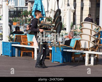 ASHKELON, ISRAEL - MARCH 04, 2022: A restaurant worker collects chairs in a street cafe. Stock Photo