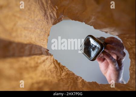 A hand tosses a black bottle into a paper bag. A mature man's hand holds the glass bottle over the open brown bag. Shot from the bottom up. Close up. Stock Photo