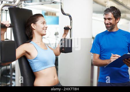 Am I doing this correctly. Cropped shot of an attractive young woman using an exercise machine while her personal trainer looks on. Stock Photo