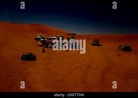 unrecognizable group of tourists and tuareg camp in Sahara Desert by night. 4X4 vehicles parked, red colored sand dunes, stars in dark sky. Stock Photo