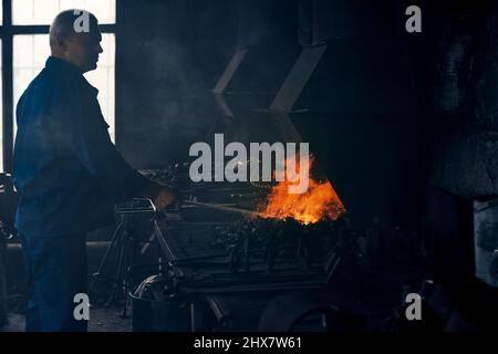 Side view of worker in dark blue suit standing near special heating equipments and working with metal in smithy. Concept of process creating modern fence.  Stock Photo