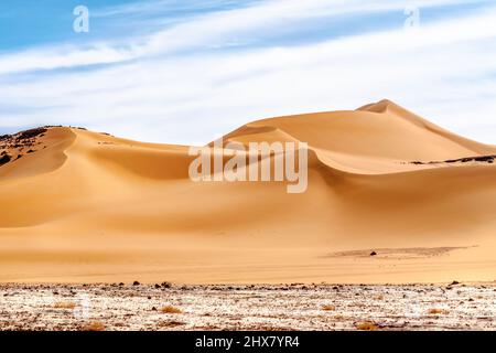 Low angle view of curvy sand dune in Sahara Desert. Colorful surface level, white reg terrain, high wavy golden colored dune, blue cloudy sky. Stock Photo