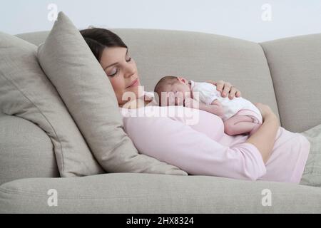 Mother resting on sofa with eyes closed and sleeping baby girl curled up on her front Stock Photo