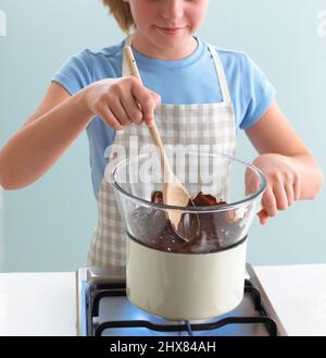 Girl using wooden spoon to stir melting chocolate in large mixing bowl on top of saucepan on gas hob, front view Stock Photo