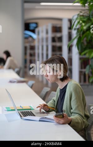Focused female university teacher using laptop while sitting in quiet cozy library Stock Photo