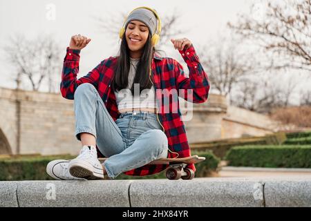 Carefree brunette woman listening to music and singing, using headphones, sitting on skateboard outdoor, copy space. Happy young lady with closed eyes Stock Photo