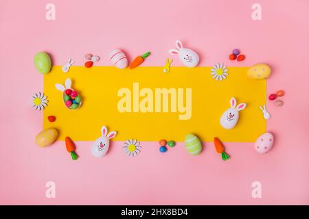 Top view festive Easter pink background with yellow frame made of colored eggs, decorative carrots, bunny rabbits, flowers, chocolate eggs. Creative b Stock Photo