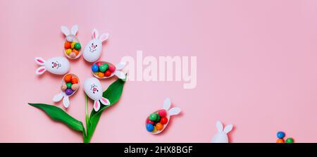 Top view Easter pink background with flower shape made of funny eggs containers with bunny ears filled with candy chocolate eggs. Bunny rabbit symbol. Stock Photo