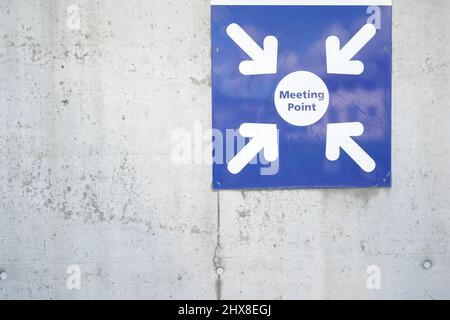 Metal shield saying meeting point attached on a concrete wall. The shield is blue and square. There are four arrows coming from the angles . Stock Photo