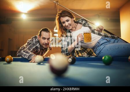Young smiling cheerful couple is playing billiards in bar and drinking beer after work. They are involved in recreational activity. Stock Photo