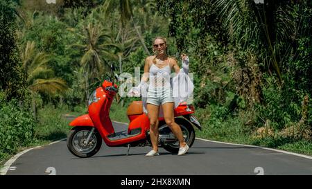 Woman on red scooter in white clothes drive on forest road trail. Dancing trip. One girl caucasian tourist in sunglasses dance, relax, rest near motor Stock Photo