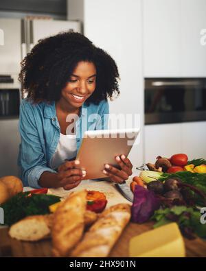 Looks like Im doing everything right. Shot of a cheerful young woman browsing on a digital tablet while being surrounded by vegetables in the kitchen Stock Photo