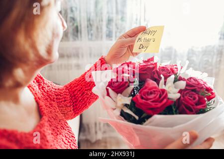 Happy Mothers Day gift. Senior woman reads greeting card on bouquet of red roses and orchids flowers at home. Present for spring holiday Stock Photo