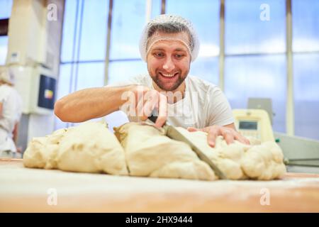Young man as a baker apprentice cuts dough with a knife in preparation for baking Stock Photo