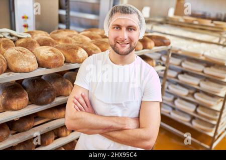 Young man as a baker apprentice with crossed arms in the bakery in front of bread shelves Stock Photo