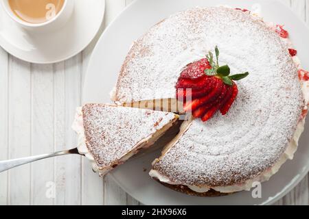 Top view of Victoria sponge cake filled with strawberries, jam and whipped cream decorated with icing sugar and strawberry on a white wooden table Stock Photo