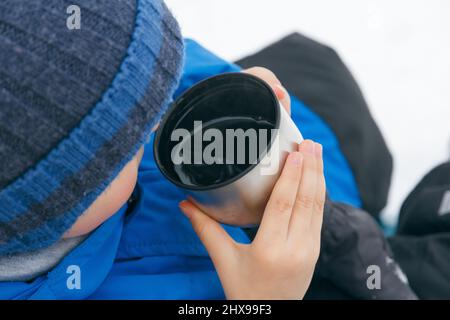 Child hands holds a cup. Child drinking a cup of tea on the street while walking. A cup of hot drink from a thermos outside in winter. Winter walk Stock Photo