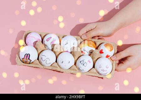 The child holds painted Easter eggs in the form of various cheerful emotions on a pink background. Yellow lights in the photo. Copy space. The concept of Happy and Peaceful Easter. Stock Photo