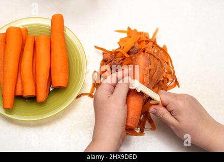 Hands of a woman peeling vegetables using a food peeler. Cleans the peel from carrots. Cooking fresh carrots before serving. Stock Photo