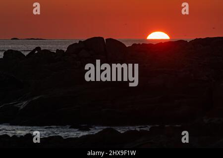 The sun hides on the horizon and behind the rocks in a reddish dusk Stock Photo