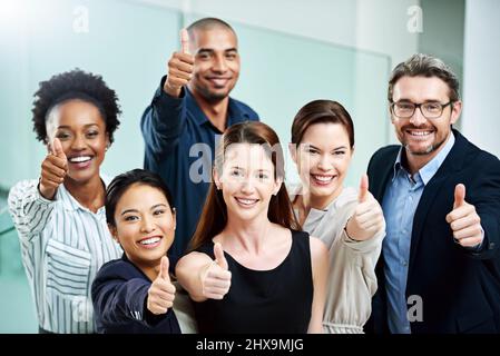 Stay positive, work hard and make it happen. High angle portrait of a group of businesspeople standing together and showing a thumbs up in the office. Stock Photo