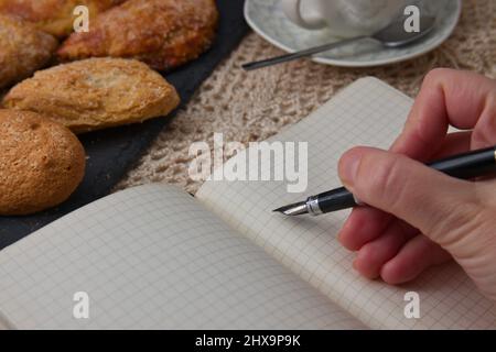 Someone is holding a pen over a notebook with an empty page next to some sweets and a cup of coffee Stock Photo