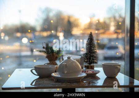 A table in a cafe with a view of the evening city. The table is set for a Christmas tea party. Garlands are burning on the window Stock Photo