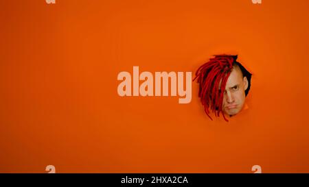 Young man with sad expression on his face sticking out of hole of orange background. Offended male with red dreadlocks purses his lips Stock Photo
