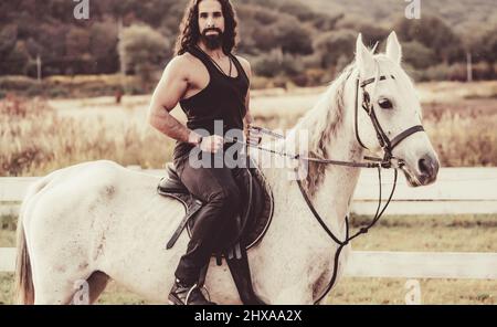 Young jockey training his horse for a ride. Good looking, hunky cowboy rides horse at beautiful country side nature. Equestrian rides his white well Stock Photo