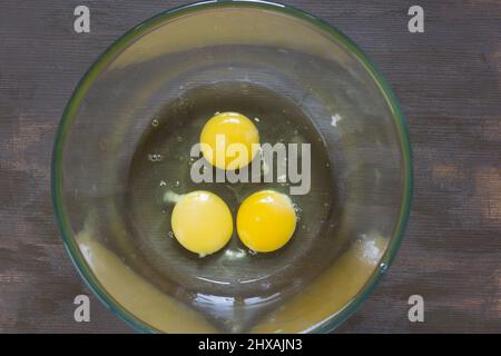 Three raw chicken yolks and whites in glass bowl on wooden table Stock Photo