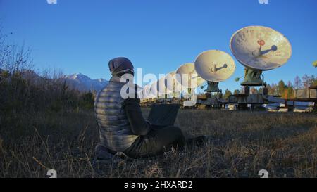 Hacker hacks the radio telescope using notebook. Man to program hacking on laptop in nature. Terrorist is hiding in camouflage balaclava. Hacker has c Stock Photo