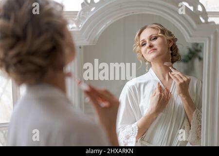 Woman massaging her face with a pink stone roller face massage at home, facial spa treatments. Caucasian girl stands in bathrobe and uses facelift kit, looks at reflection in mirror. Skin care at home Stock Photo