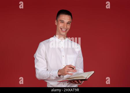 Photo an attractive business thinking guy holding planner thinking about idea of a new startup project in a white shirt and light trousers, isolated dark red color background Stock Photo