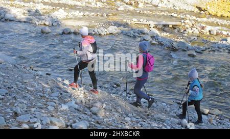 Woman do Nordic walking in nature near mountain river. Girls and children use trekking sticks and nordic poles, backpacks. Family travels sports. Kid Stock Photo