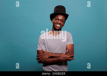 African american man with arms crossed wearing top hat to create magical artistic performance, showing confidence for vintage fashion. Young adult with theater prop costume in studio. Stock Photo